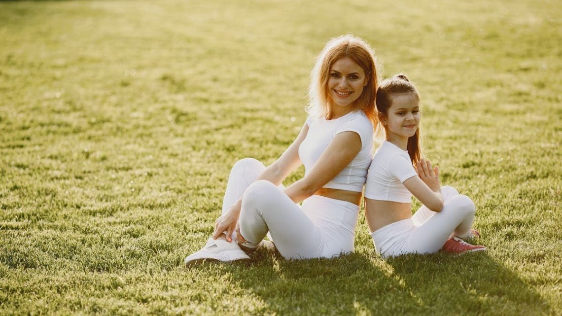 A mother and daughter sit back to back in matching activewear on a sunny grass field, enjoying a summer day together.